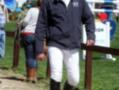 Un hombre vestido con una chaqueta negra de marca, pantalones blancos y botas negras camina por un camino asfaltado. En el fondo, hay una persona con una chaqueta blanca y pantalones azules. La imagen parece ser de un evento equestre, posiblemente una competencia o reunión.