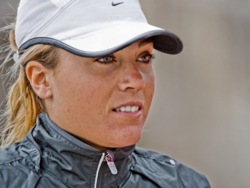 A una mujer con cabello largo y rubio, llevando un gorro de béisbol blanco y gris, una chaqueta deportiva azul y una correa roja en la parte superior. La escena parece ser al aire libre, posiblemente en un campo de béisbol o un parque.