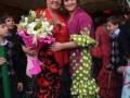 Dos mujeres en trajes flamencos posan sonrientes, rodeadas por niños y adultos. La escena parece ser una fiesta o evento cultural español con decoración típica y vestimenta tradicional.