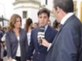Un hombre con micrófono de televisión entrevista a una mujer en un evento formal. La escena se desarrolla frente a un edificio histórico con balcones y una granja en el fondo.