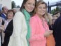 Dos mujeres jóvenes posan juntas en una celebración. La de la izquierda lleva un abrigo blanco con un pañuelo verde, y la de la derecha usa una chaqueta rosa. Ambas sonríen para la cámara, rodeadas de personas en un entorno público con una estructura metálica visible.