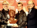 Tres hombres sonrientes posan con un busto de bronce en una alfombra roja. El medio sostiene el busto mientras los otros dos lo miran con orgullo. En el fondo, un cartel con la palabra "ARTISTAS" y una insignia de la Asociación de Periodistas de Arte.