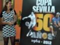 Una mujer sonríe mientras camina frente a un cartel que anuncia la Copa Sevilla, celebrando sus 50 años desde 1962 hasta 2012.