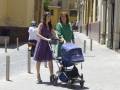 Dos mujeres caminan por una calle adoquinada empujando un cochecito de bebé, con edificios de fachadas amarillas al fondo.