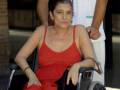 Una mujer en silla de ruedas con un vestido rojo, acompañada por una persona que la sostiene.