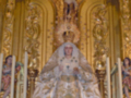 Imagen de una Virgen María vestida con un manto dorado, rodeada por detalles artísticos y figuras religiosas en una iglesia.