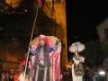 Imagen nocturna de una calle con personajes vestidos de manera antigua, posiblemente durante una celebración o fiesta. La estructura histórica en el fondo sugiere un entorno europeo, posiblemente de España. La escena está iluminada por farolas y luces, con un cielo oscuro que sugiere una noche de invierno.