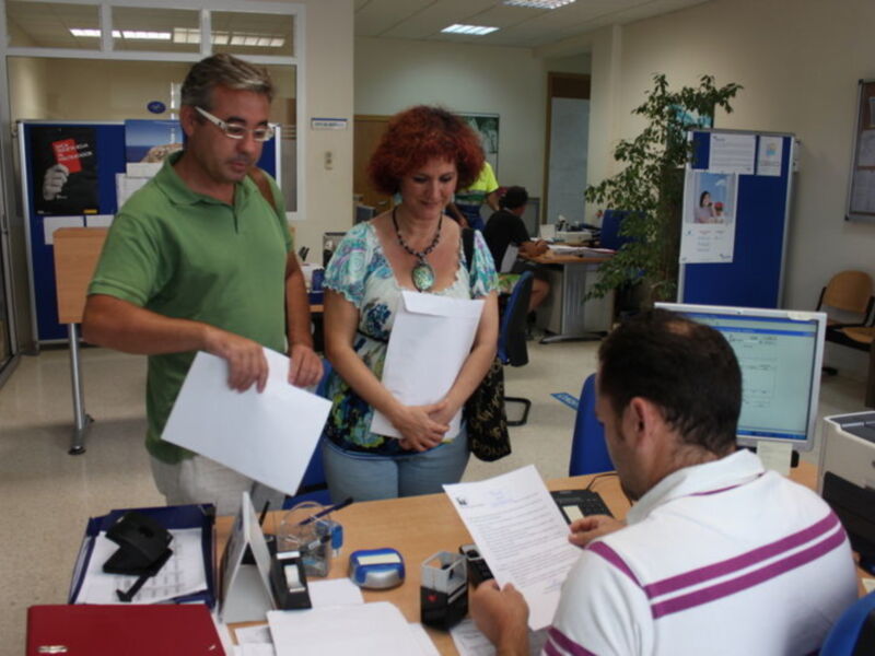 En una oficina, un hombre y una mujer conversan mientras otro trabaja en un escritorio. La imagen muestra documentos, una computadora y una planta en el fondo.