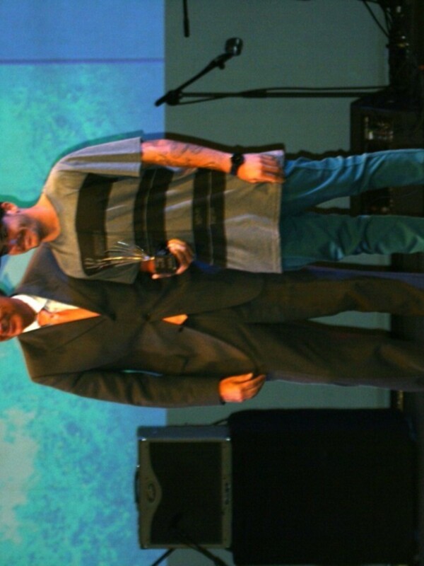 Dos hombres, uno en traje y otro en camiseta con pantalones azules, posan juntos en un escenario. El hombre en traje sostiene una copa mientras el otro mira hacia la cámara.