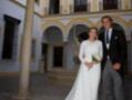 Una pareja casada posa en un patio con arcos y columnas, el hombre lleva traje negro y la mujer una vestido blanco largo con un ramo de flores.