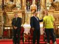 En una iglesia ornamental, tres personas posan frente a un altar dorado con flores y velas. La imagen muestra detalles de la arquitectura barroca, con estatuas y adornos dorados.