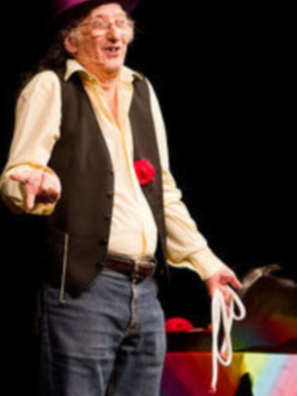 Un hombre con sombrero púrpura y corbata roja, posiblemente un mago o payaso, está en una escena teatral con un fondo negro y un arco iris.