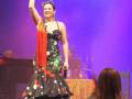 Una mujer en un vestido flamenco negro con lunares rojos y una falda larga, levanta el brazo derecho en un gesto de triunfo. La escena está iluminada con una luz azul y el público asiste a la actuación.