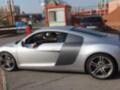 Un conductor maneja un Audi R8 en una calle urbana, destacando su diseño deportivo y color plateado.
