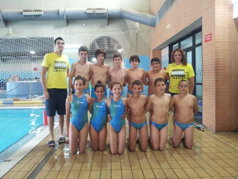 Un grupo de niños y niñas posando en una piscina interior. Los niños están vestidos con trajes de baño azules y los niñas con trajes de baño verdes. Al fondo, se pueden ver tuberías y una pared de ladrillos.
