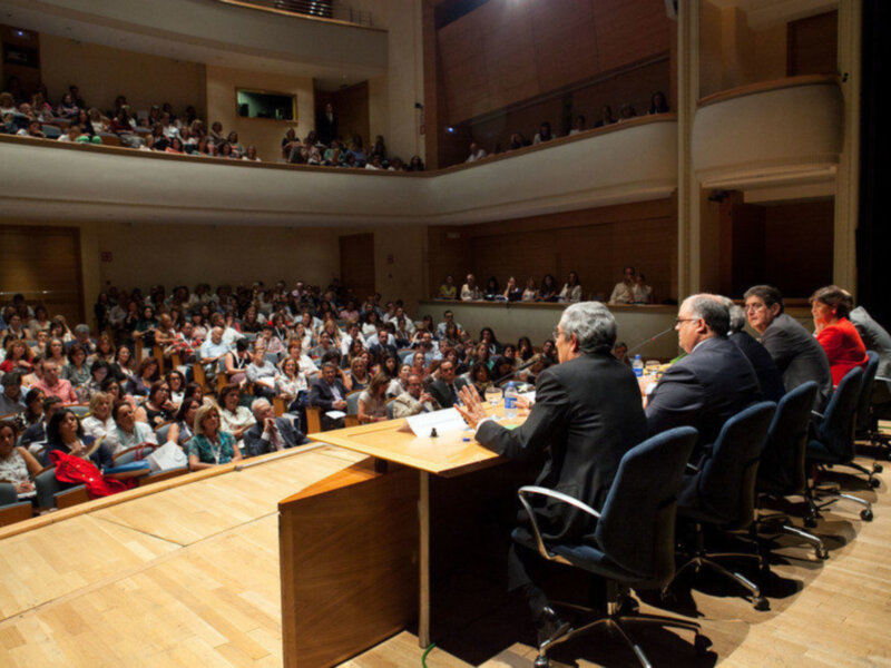 Una audiencia numerosa en un auditorio, con una mesa central y varios asistentes sentados en sillas giratorias. La imagen muestra a una persona hablando frente a la audiencia, con un fondo de madera y luces iluminando el lugar.