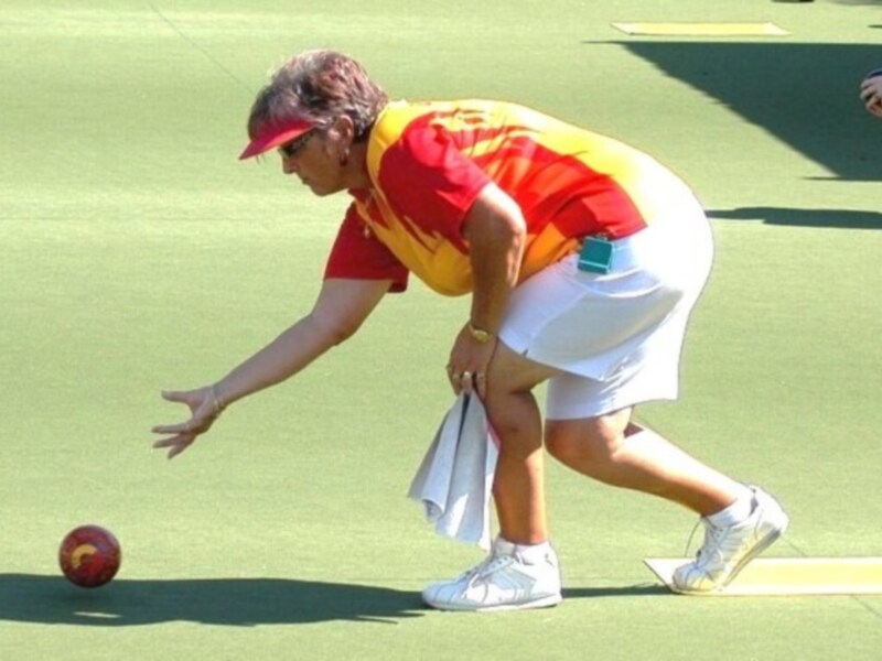 Una persona vestida con una camiseta roja y amarilla, pantalones cortos blancos y zapatillas deportivas está en una cancha de bolo. La persona está en una postura de lanzamiento, con el pie derecho levantado y la mano izquierda extendida hacia un bolo rojo en el suelo. La imagen muestra una escena deportiva, probablemente un partido de bolo.