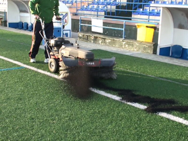 Un trabajador de la construcción está utilizando una máquina para pulir el césped artificial en un estadio. La máquina tiene una gran brocha que está raspando el césped, dejando un patrón de líneas en la superficie. La escena se desarrolla en una zona de césped artificial, con un fondo que incluye una estructura blanca y azul del estadio.