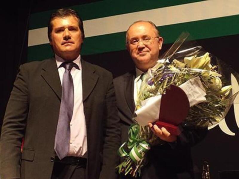 Dos hombres en trajes posan con un hombre que sostiene una corona y flores. El fondo muestra un escudo nacional y la palabra "ALU".