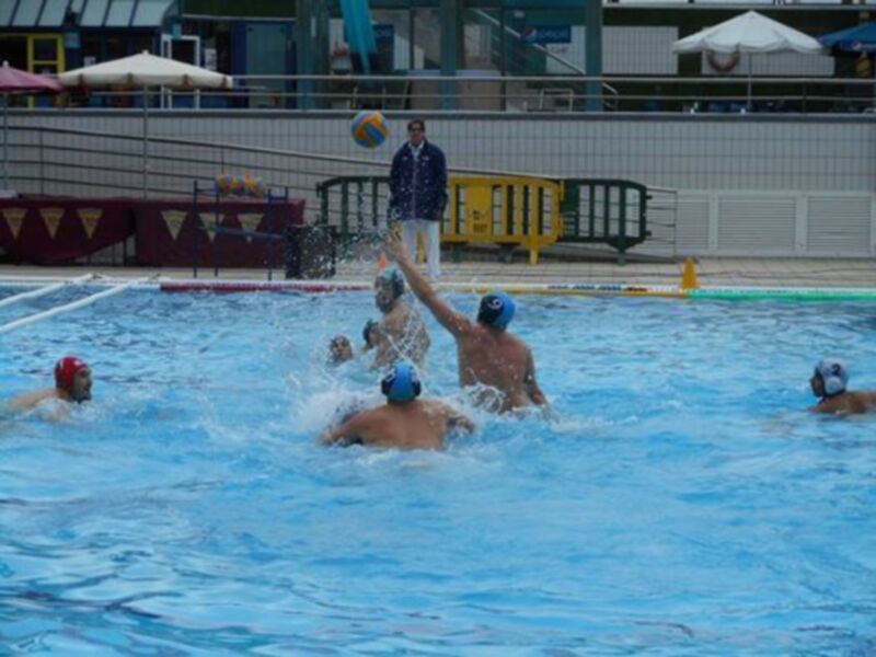 Jugadores de waterpolo en acción en una piscina. Un árbitro observa el juego desde la orilla. La imagen muestra un ambiente deportivo activo y competitivo.