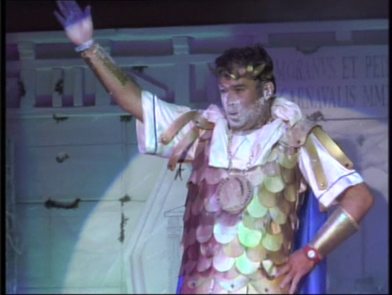 Un hombre vestido con un traje de gladiador antiguo, incluyendo una armadura de escamas y un tocado con cuernos. Está en una escena teatral o de espectáculo, posiblemente representando a un personaje histórico o mitológico.