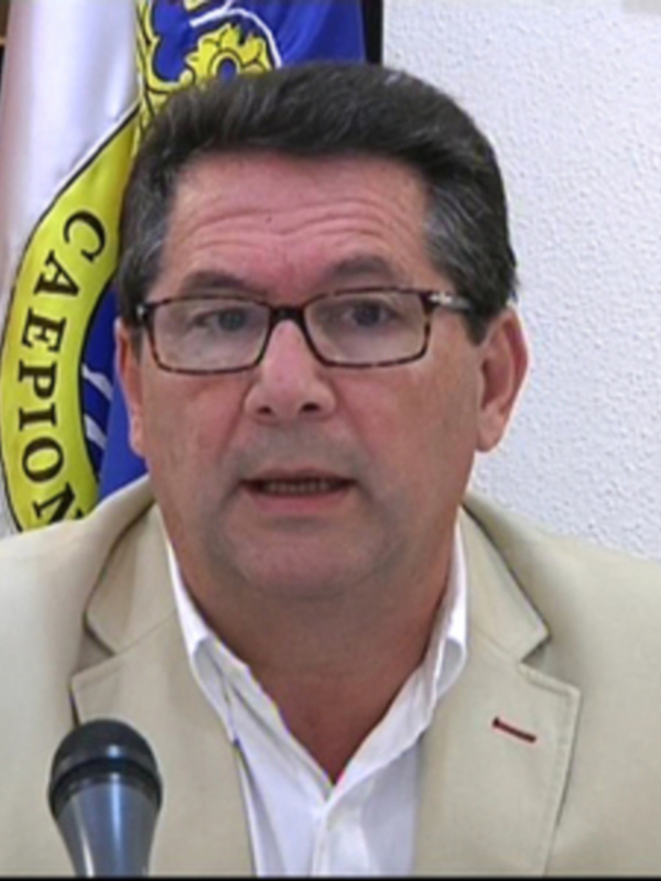 El pié de foto es un hombre con cabello corto y barba, lleva gafas y una chaqueta beige. Está frente a un fondo con banderas, una de las cuales tiene el escudo de la ciudad. La imagen parece ser de un evento oficial o conferencia, ya que el hombre está hablando en un micrófono.