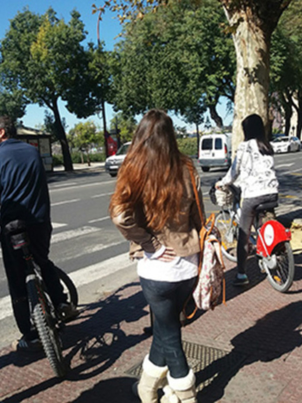 En un entorno urbano, dos personas caminan por la acera mientras otra se detiene junto a su bicicleta. El paisaje está lleno de árboles y edificios, con un cielo azul claro. La escena transmite una sensación de tranquilidad y actividad cotidiana en un entorno urbano.