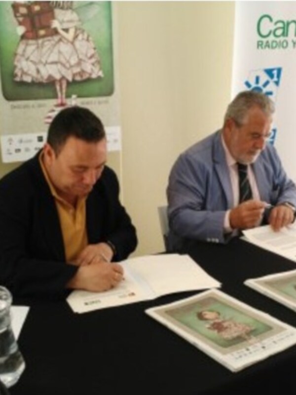 Dos hombres firman documentos en una mesa con carteles de Canal Sur Radio y Televisión en el fondo.