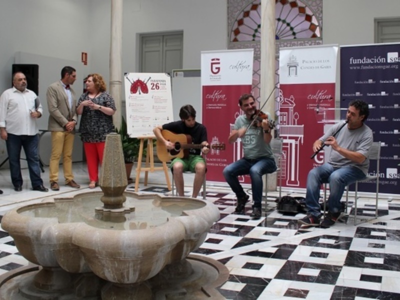 En una sala con suelo de baldosas negras y blancas, un grupo de personas observa a tres músicos sentados en sillas. Los músicos tocan guitarra y bajo eléctrico frente a carteles promocionales de Fundación Telefónica. Una fuente de piedra se encuentra en el centro, rodeada por los asistentes.