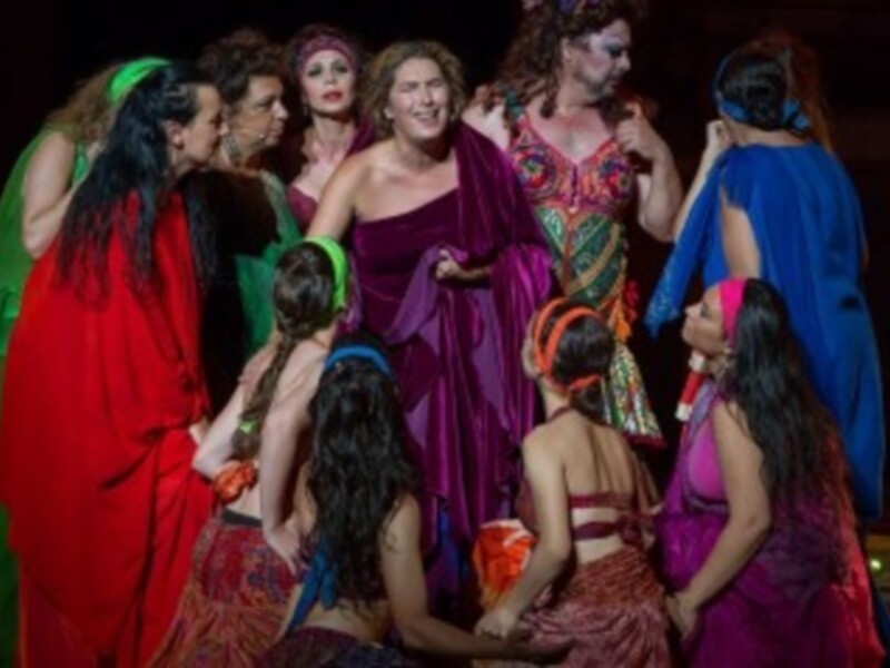 Una escena teatral con varias actrices vestidas de colores vibrantes, posiblemente en un drama histórico o mítico. La iluminación es dramática, enfocándose en los rostros y expresiones de las actrices.