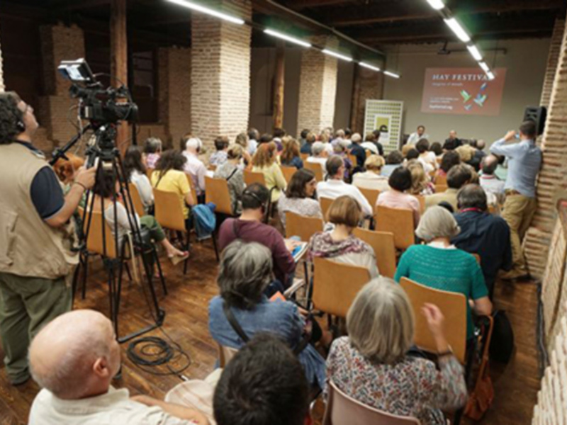 Una sala llena de personas sentadas en sillas, mirando hacia adelante. En el centro hay una pantalla con un cartel que dice "May Festival". A la izquierda, un hombre está filmando a través de una cámara. La pared detrás de las personas es de ladrillo y tiene luces de techo.