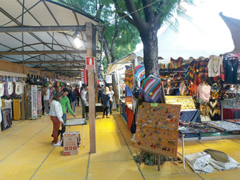 Mercado de artesanías en un día soleado. Varios stands con productos coloridos y artesanales. Suelo amarillo, toldos de metal y árboles en el fondo.
