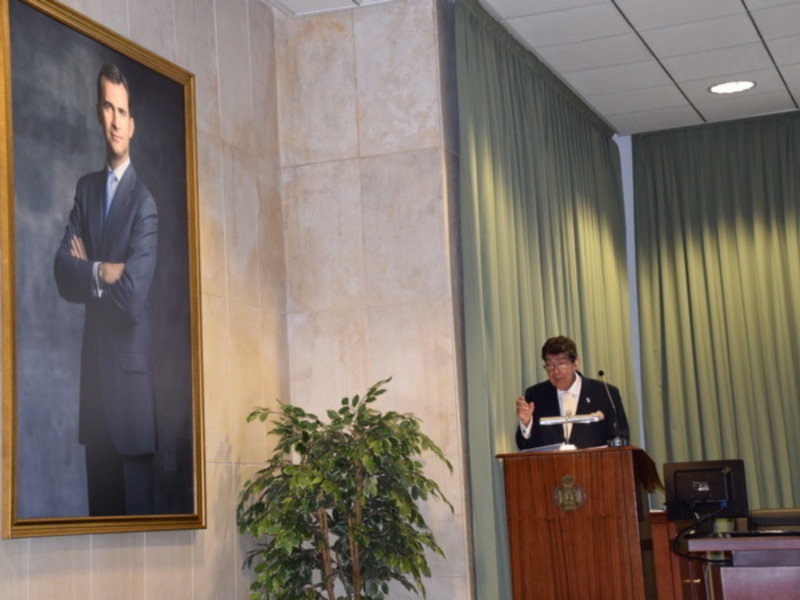 Un hombre en una sala de conferencias, con un retrato de un hombre en la pared y una planta en el suelo.
