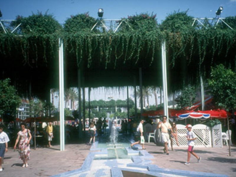 Un grupo de personas disfrutando en un parque público con una fuente central, rodeada por árboles y toldos. La escena es soleada con un cielo azul claro, y varias personas están caminando o sentadas cerca de la fuente.
