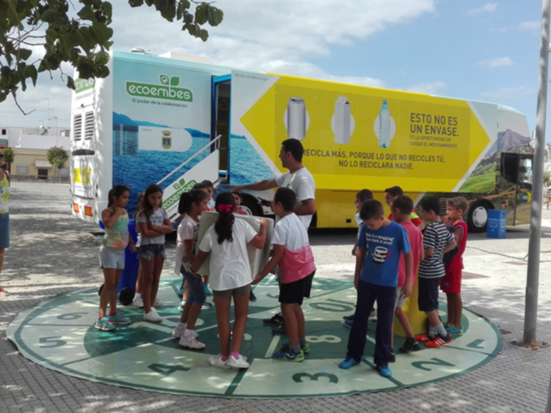 Un grupo de niños y adultos observan un camión de reciclaje con el lema "Recicla más, porque los desechos no son envases". La imagen muestra un ambiente educativo y ambiental.