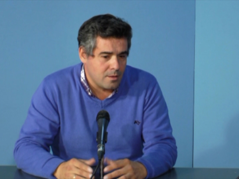El hombre está sentado en una mesa con un fondo azul claro, hablando al micrófono.