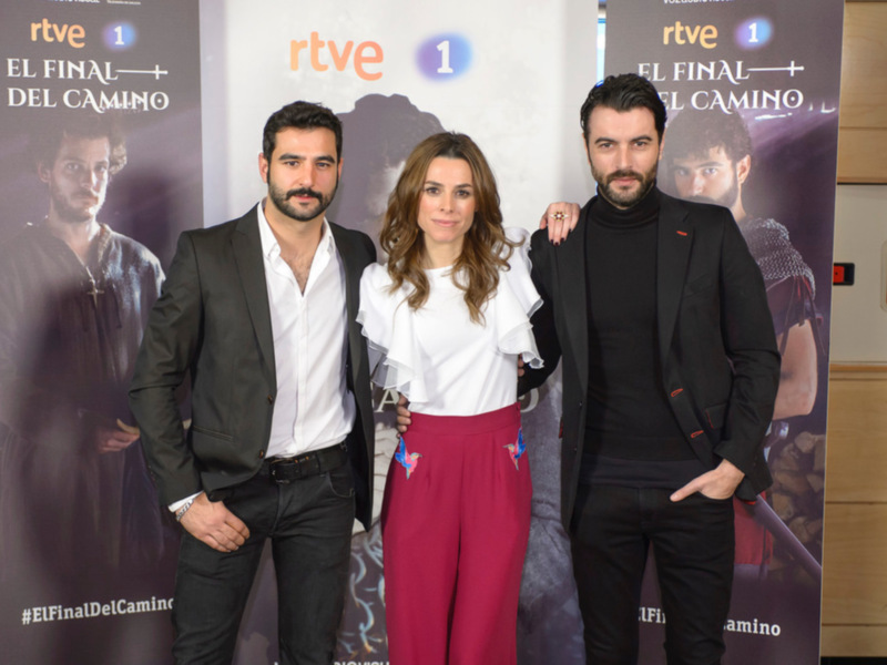 Se ven tres personas posando frente a un póster de la serie "El Final del Camino" emitida por RTVE. El póster muestra a dos personajes principales en una escena dramática, con el nombre de la serie y el logo de RTVE. La persona central lleva una blusa blanca y pantalones rojos, mientras que las otras dos lucen trajes formales.