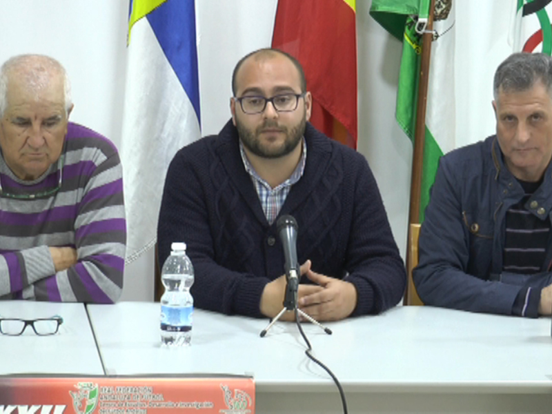 Tres personas sentadas en una mesa con banderas de diferentes países al fondo. La imagen parece ser de un evento oficial o conferencia, con una botella de agua y un micrófono en la mesa.