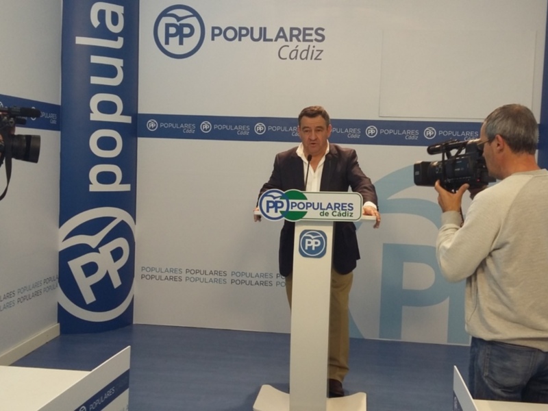 Un hombre habla en un escenario con banderas y logotipos de PP Populares. Un hombre filmando a la izquierda, otro con una camiseta blanca y pantalones azules en el fondo.