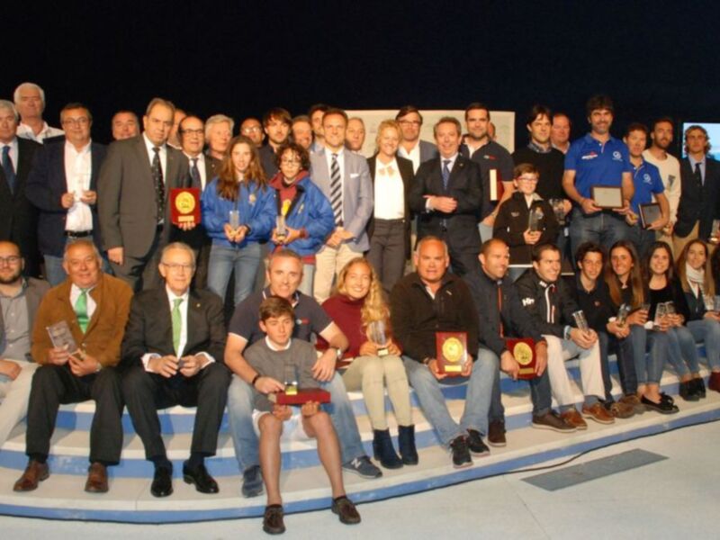 Una gran imagen de un grupo de personas posando juntos en una escena formal. La mayoría están vestidos con trajes y chaquetas, algunos con medallas o trofeos en sus manos. La escena parece ser un evento importante, posiblemente una conmemoración o ceremonia.