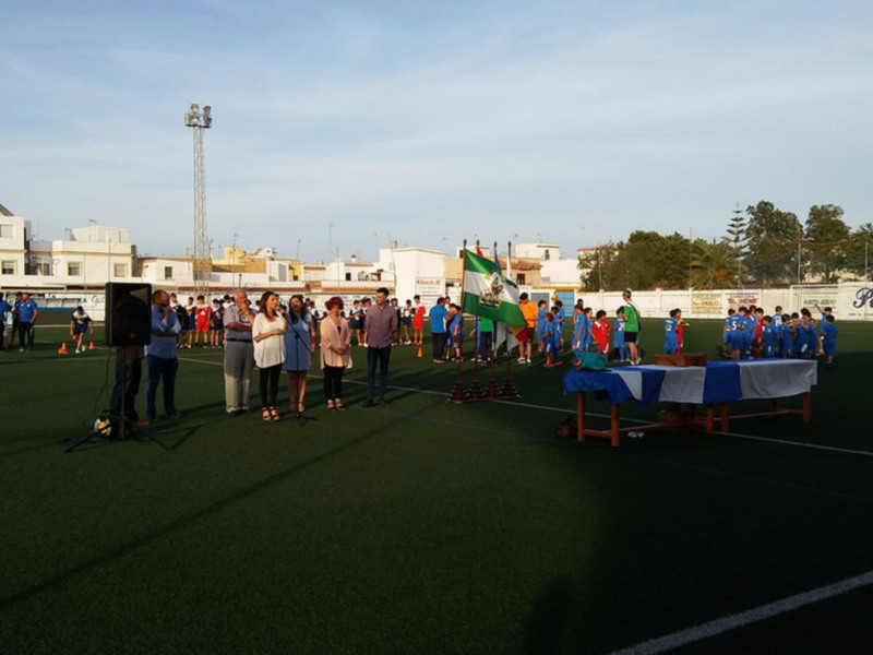 Un equipo de fútbol se prepara para un partido en un campo de fútbol artificial. Los jugadores están vestidos con camisetas y pantalones de color azul, mientras que los entrenadores y miembros del cuerpo técnico están vestidos con camisas más oscuras. En el fondo, se pueden ver edificios y árboles a lo lejos bajo un cielo azul claro.