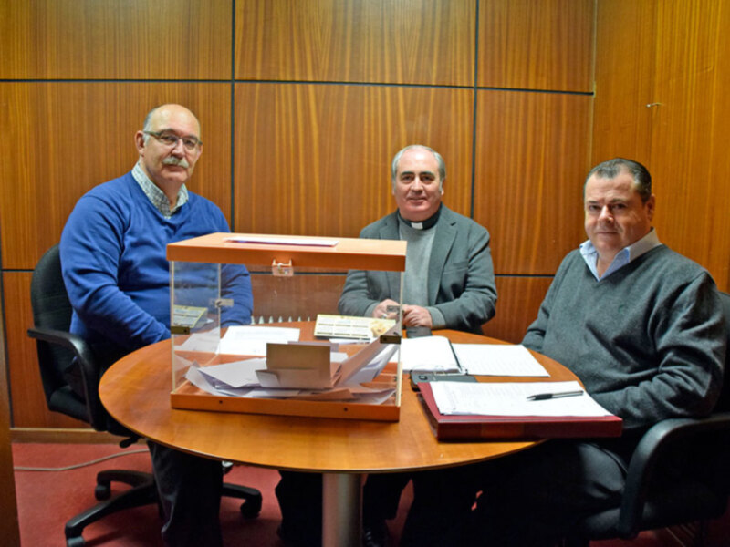 Tres personas sentadas en una mesa de madera, con documentos y un portafolios en el centro. La pared detrás es de madera, y hay una silla vacía a la izquierda.