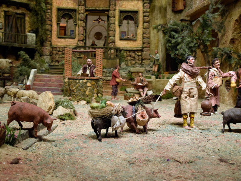 Un escenario de pesebre navideño con figuras humanas y animales, incluyendo un hombre con una lanza, una mujer con un cesto de leña y varios animales como cerdos y ovejas. La escena está ambientada en un entorno rural con edificios de piedra y una fuente.