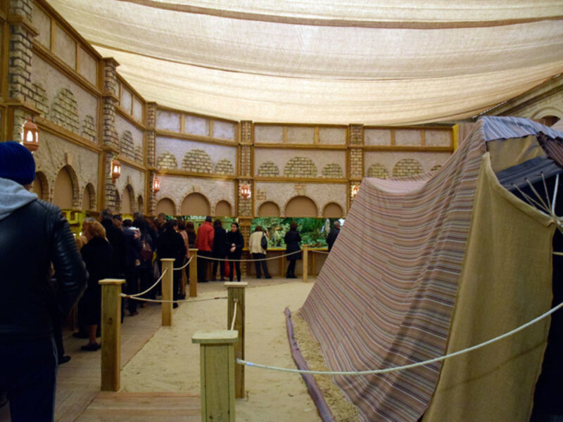 Interior de una sala con techo curvo y paredes decoradas con motivos geométricos. Personas observando objetos en vitrinas. Cubierta de tela con rayas multicolores y estructura metálica en el fondo.