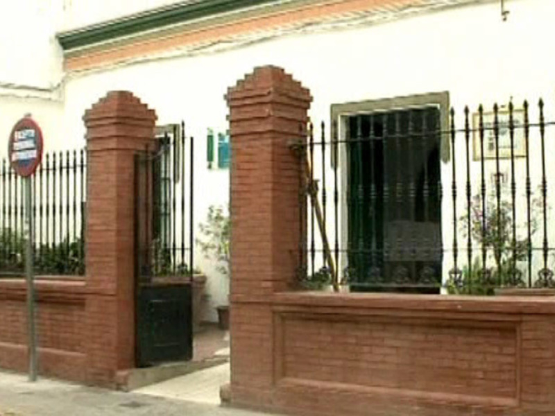 Fachada de una casa con una cerca de hierro forjado y un cartel rojo en la esquina. La casa tiene ventanas con persianas negras y un pequeño jardín con plantas. La cerca está en buen estado, y la casa parece ser de estilo colonial.
