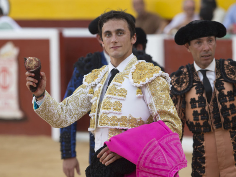 Un torero sostiene un capote de color rosa y blanco, mientras otro torero con traje tradicional espera en el fondo. La imagen se desarrolla en una plaza de toros con un público observando la escena.
