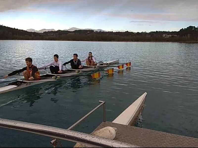 Cuatro personas remando en un bote de remo en un lago tranquilo con montañas y nubes en el cielo.