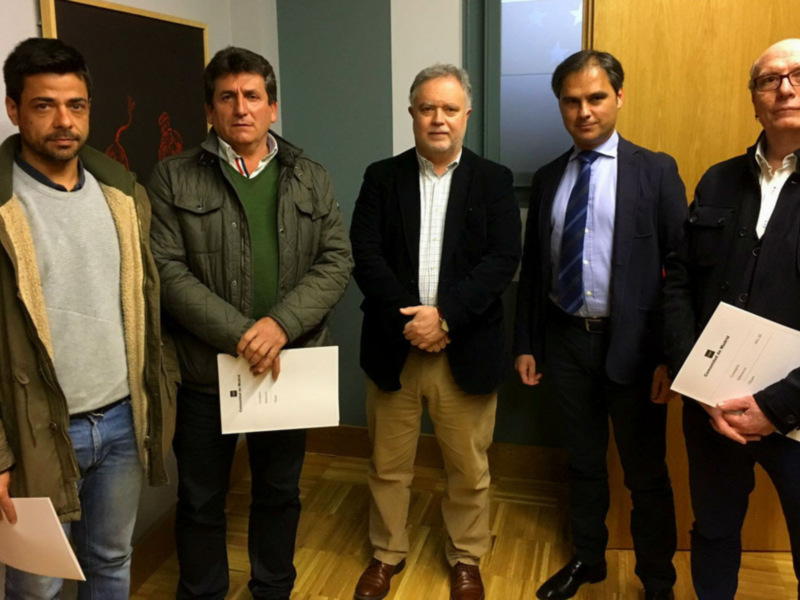 Grupo de personas en una reunión formal, posiblemente en un entorno de oficina o sala de conferencias. Todos están vestidos formalmente y algunos sostienen documentos, lo que sugiere una reunión de trabajo o presentación. El ambiente es profesional y ordenado, con paredes de color claro y muebles de oficina.