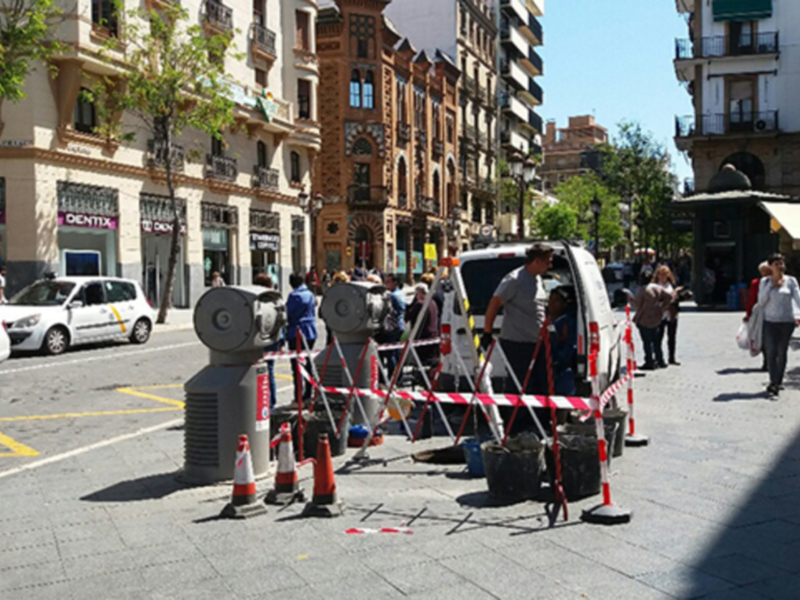 Se observa una calle de una ciudad con edificios históricos y modernos. Hay varios vehículos estacionados, incluyendo un taxi amarillo con patrón blanco. También se pueden ver personas caminando y un par de casas con balcones. La calle está bordeada por árboles y hay una señal de tráfico con un reloj en el centro. El cielo es claro y azul, lo que sugiere un día soleado.