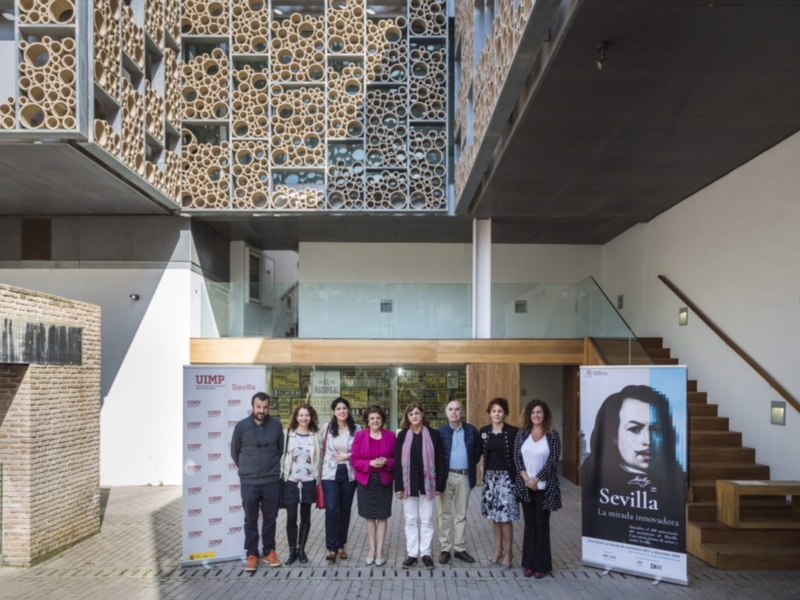 Fotografía de un grupo de personas frente a una estructura moderna con paneles de madera y piedra. En el fondo, se puede ver una escalera y un póster con la imagen de una mujer y la palabra "Sevilla".